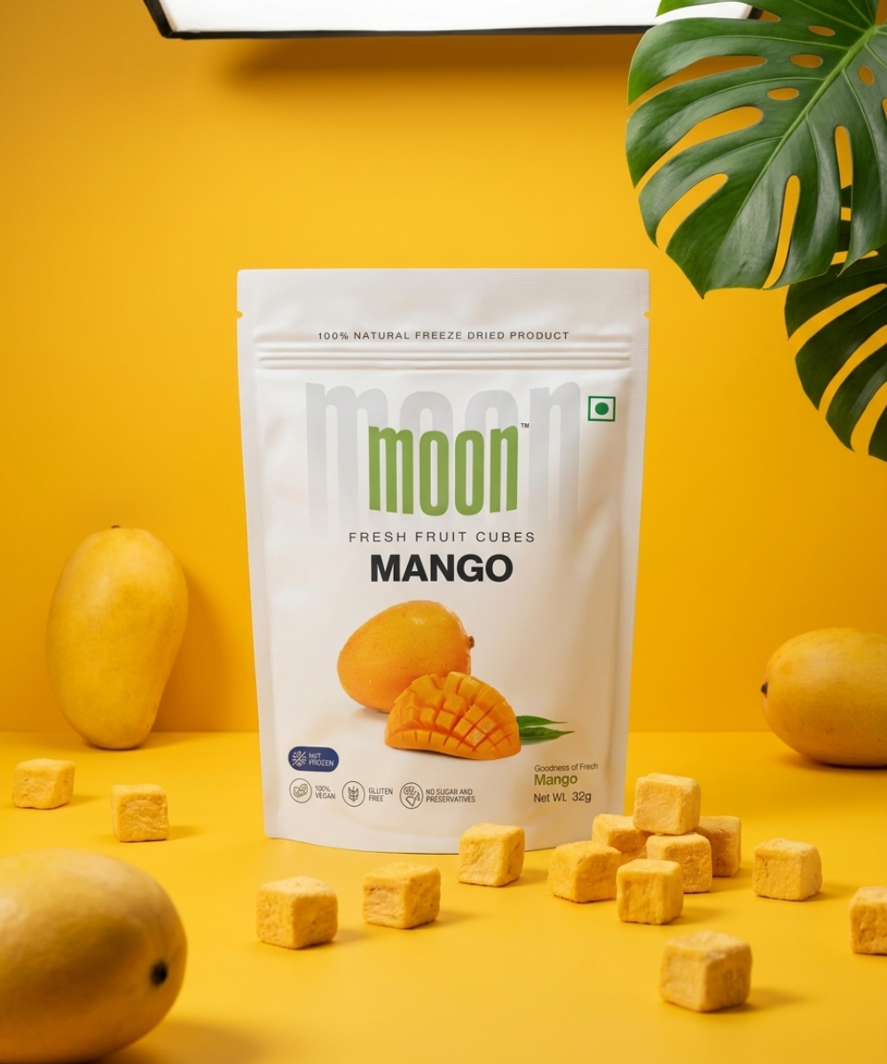 A white packet of Themoonstoreindiaβs Moon Freeze Dried Mango Cubes stands upright on a yellow surface with fresh mangoes and cubes around itβa delicious, healthy snack with no added sugar.