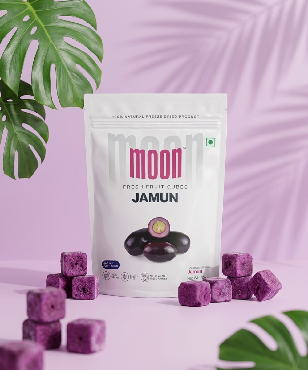 A white packet of "Moon Freeze Dried Jamun Cubes" by Themoonstoreindia stands upright with freeze-dried jamun chips scattered nearby, set against a pink background with green leavesβa tasty, gluten-free treat for natural sugar cravings.