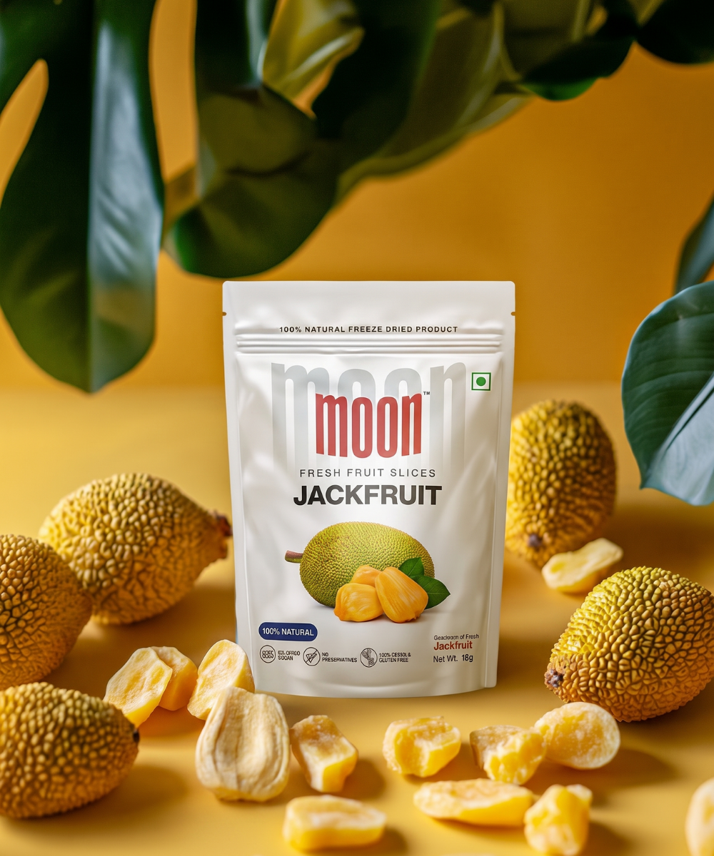 A pack of Themoonstoreindia's Moon Freeze Dried Jackfruit stands upright among fresh jackfruits and loose chips on a yellow surface, with green leaves behindβoffering a 100% natural, healthy snack choice.