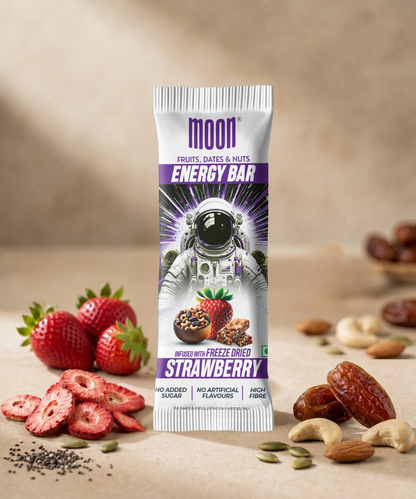 A MOON Energy Bar - Strawberry by MOONFREEZE FOODS PRIVATE LIMITED stands upright, surrounded by freeze-dried strawberries, dates, nuts, and seeds on a surface with a blurred neutral background.