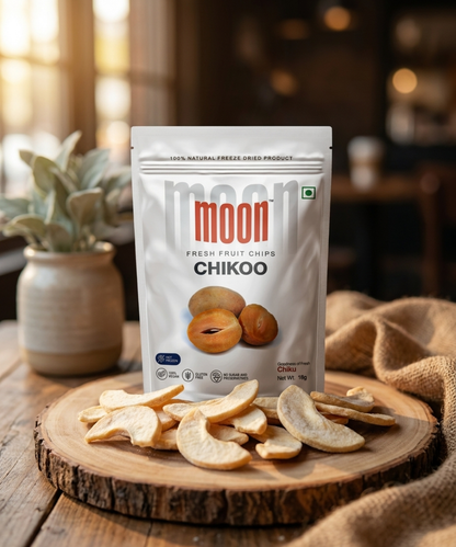 A bag of Themoonstoreindia’s Moon Freeze Dried Chikoo stands on a wooden table, with 100% natural dried fruit chips on a plate in front and a potted plant behind—perfect as an energy-boosting snack.