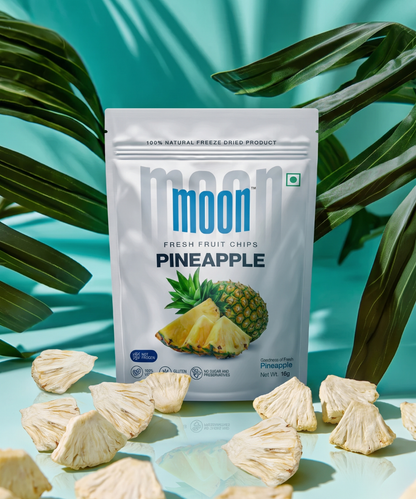 A packet of Moon Freeze Dried Pineapple by Themoonstoreindia stands upright among dried pineapple pieces with tropical leaves and a teal background—an irresistibly healthy snack containing no added sugar.